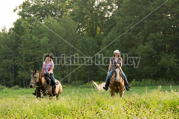 Two young women horseback riding western through summer pasture fields.