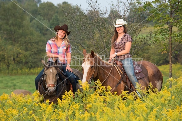 Two young women horseback riding western through summer pasture fields.