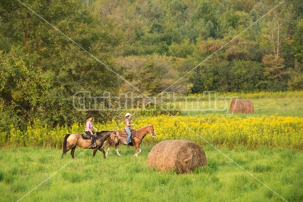 Two young women horseback riding western through summer pasture fields.