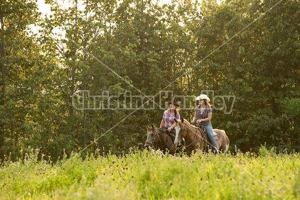 Two young women horseback riding western through summer pasture fields.