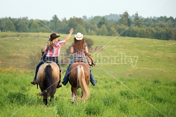 Two young woman horseback riding western goofing around stealing each others cowboy hats.