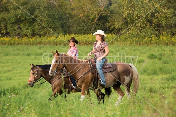 Two young women horseback riding western through summer pasture fields.